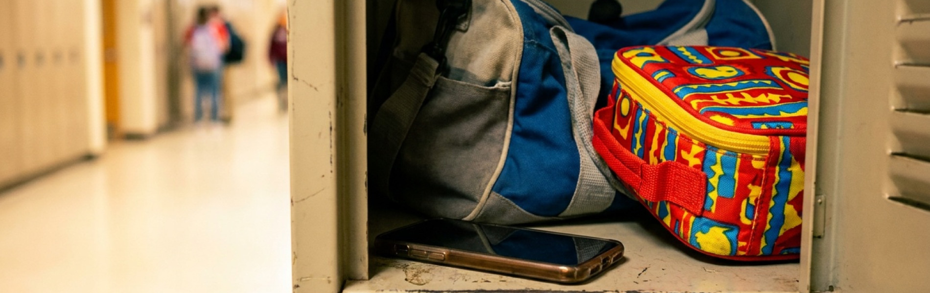 Een smartphone met het scherm naar beneden in een open schoolkluisje, naast een gymtas en een broodtrommel